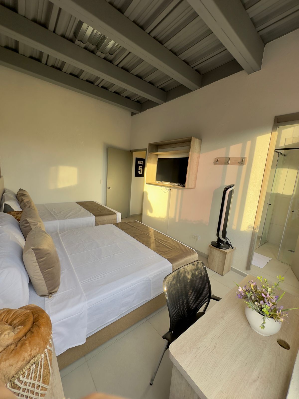 A modern hotel room with two beds, a desk with a chair, a TV mounted on the wall, and a shower area.