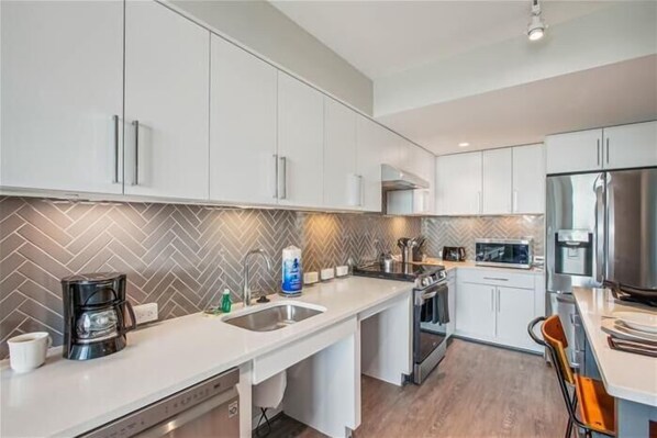 Una cocina moderna con armarios blancos, salpicadero con estampado de espiga, electrodomésticos de acero inoxidable y una isla central con pileta.