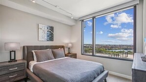 Un dormitorio moderno con una cama grande, mesitas de noche, un escritorio y un amplio ventanal con vistas a la ciudad.