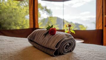 Una toalla doblada con un rosal sobre la cama, una ventana con vistas a la vegetación y montañas, y cortinas.