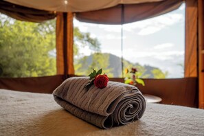 A neatly folded towel with a rose on a bed, a window with a view of greenery and mountains, and curtains.