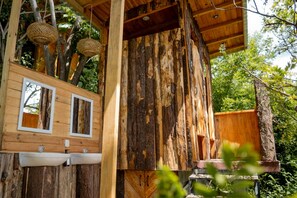 A rustic wooden cabin with two white-framed windows, a wooden bench, and hanging woven baskets.