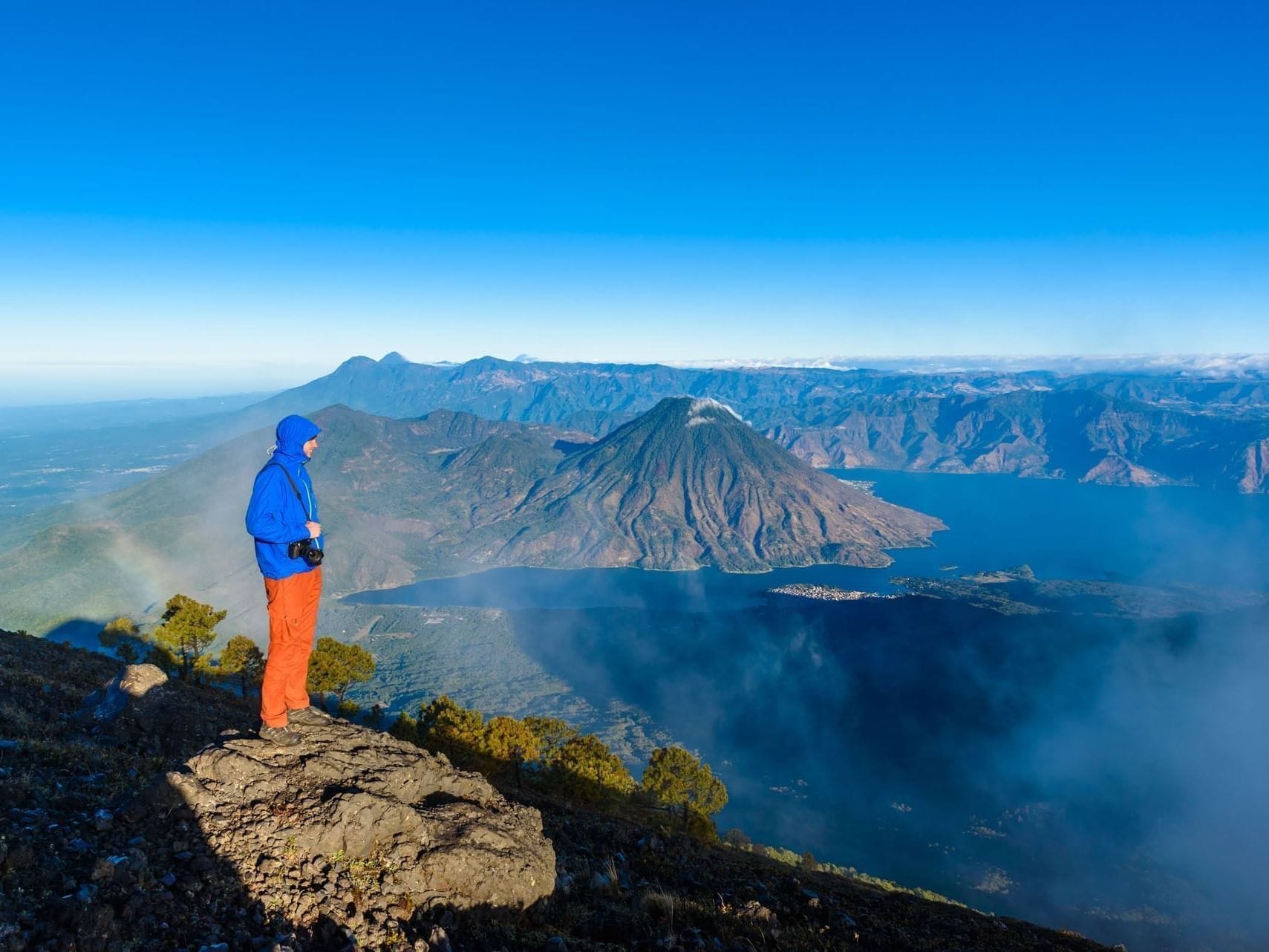 A hiker on a rocky outcrop overlooking a mountainous landscape with a lake and distant mountains.