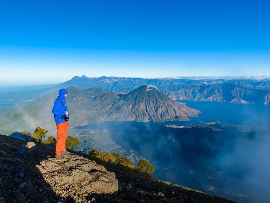 A hiker on a rocky outcrop overlooking a mountainous landscape with a lake and distant mountains.