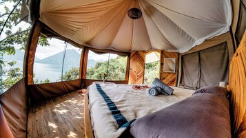 Una tienda de campaña acogedora con una cama, suelo de madera y vistas a un lago y montañas.