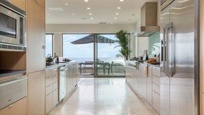 A modern kitchen with a view of the ocean, featuring stainless steel appliances, wooden cabinets, and a dining area with a table and chairs. - Jewel by Avantstay Panoramic Ocean Views in La Jolla! (La Jolla)