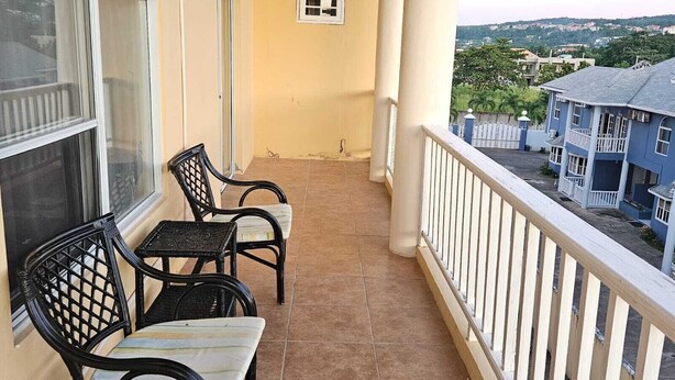 Un balcon avec deux fauteuils en osier et une petite table, donnant sur un quartier résidentiel avec des maisons et de la verdure.