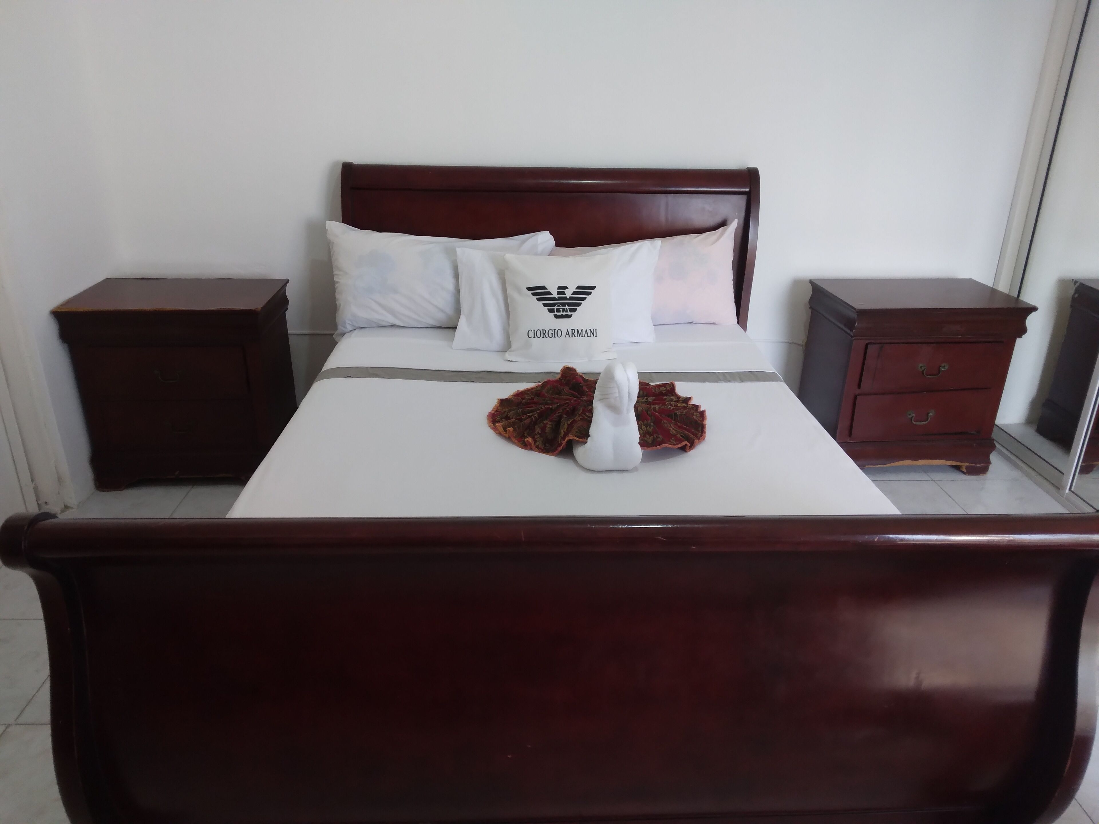 Una cama bien hecha con sábana blanca, dos almohadas y un adorno sobre la cama. Hay dos mesitas de noche de madera, una a cada lado de la cama.