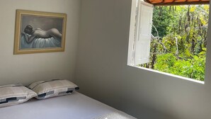 A bedroom with a bed, a hanging light fixture, an air conditioning unit, a window with a view of greenery, and a framed painting on the wall. - Recanto Minduba (Japaratinga)
