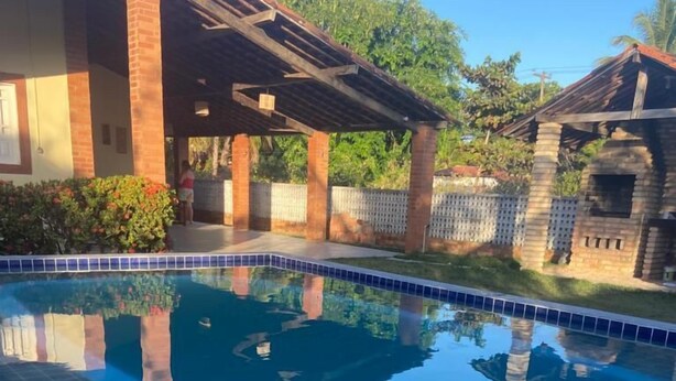 A swimming pool with clear blue water, surrounded by a brick deck and a covered patio area. - Recanto Minduba (Japaratinga)