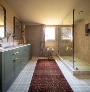 Un baño con un espejo grande, un tocador con lavamanos, una zona de ducha y una alfombra estampada.