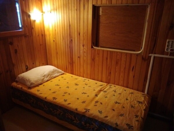 Un lit simple avec une couvre-lit à motifs floraux jaunes et un oreiller blanc, dans une pièce aux murs en bois et dotée d’une petite fenêtre.