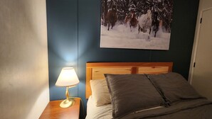 Ein Schlafzimmer mit einem hölzernen Kopfende, einer Nachttischlampe, einem Bett mit grauer Bettwäsche und einem Gemälde von Pferden in einer verschneiten Winterlandschaft an der Wand.