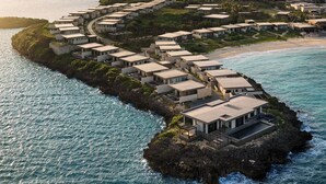 Un complejo turístico costero con villas dispuestas a lo largo de una costa curva, conectadas por senderos, con vistas al mar y un paisaje distante.
