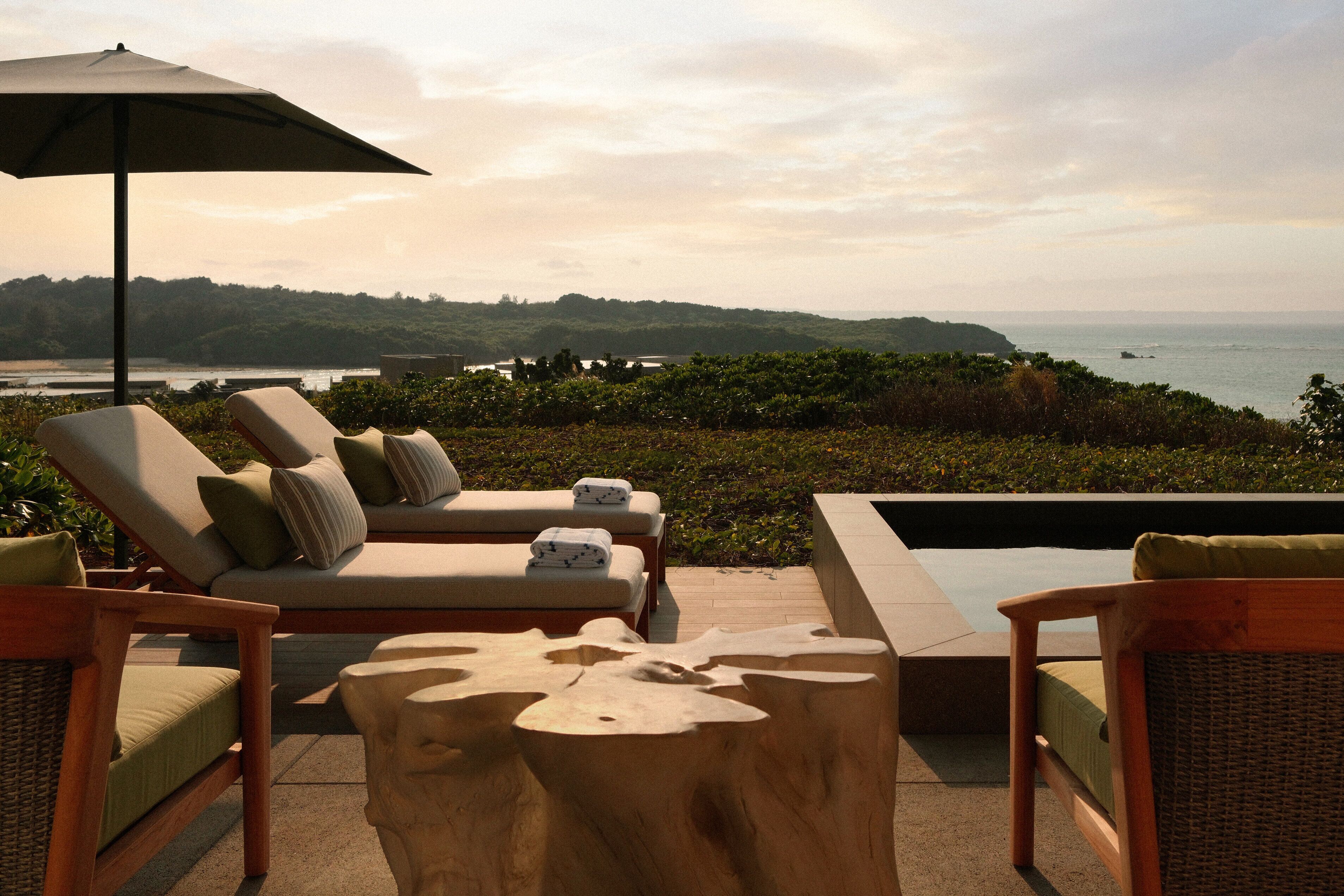 A sunlit outdoor seating area with lounge chairs and a wooden table, overlooking a coastal landscape.