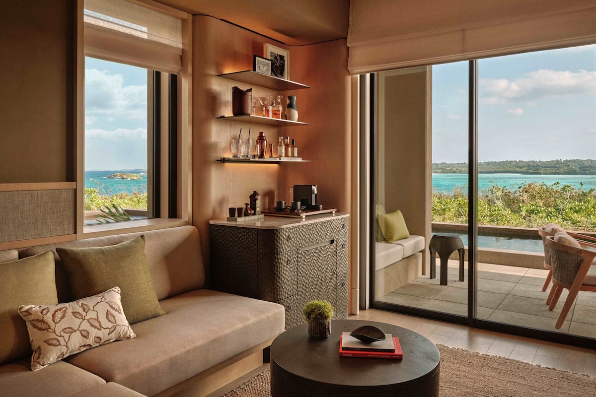 Una sala de estar moderna con un sofá, una mesa de centro y estanterías decoradas. Un amplio ventanal corredera da a un balcón con vistas al océano.