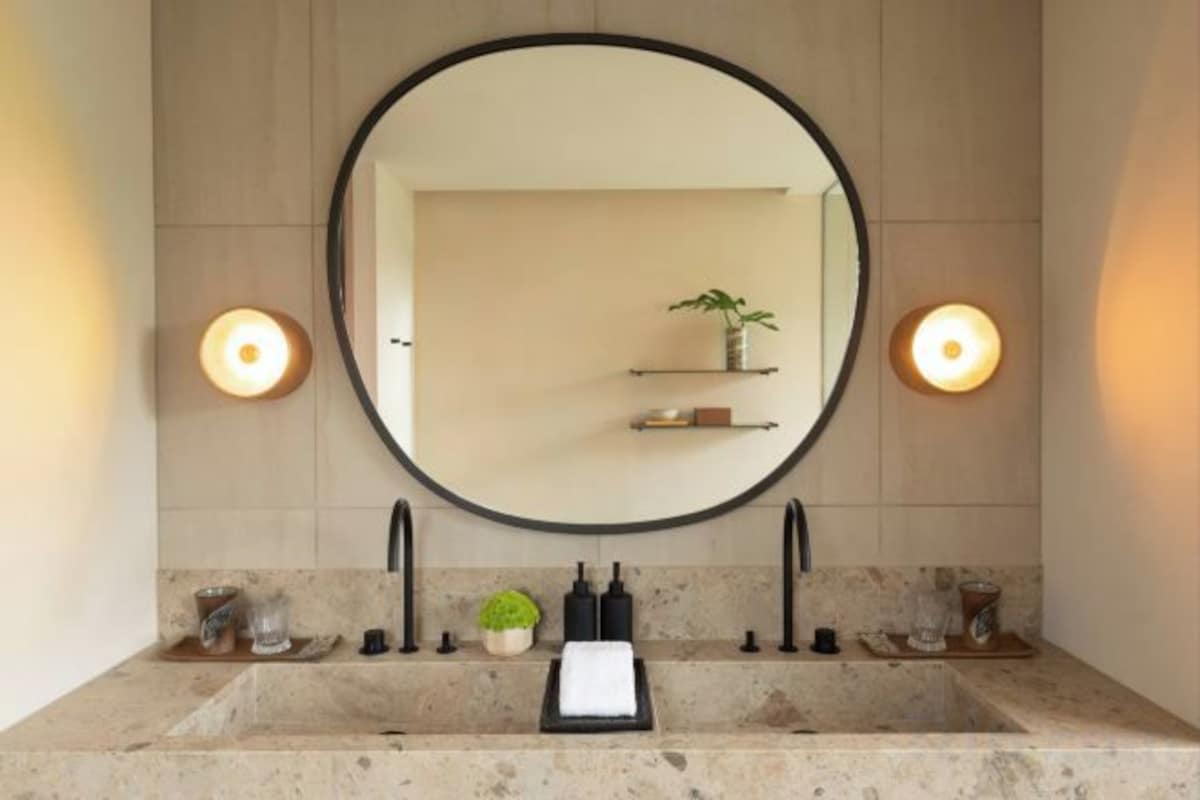 A bathroom with a large round mirror, two wall-mounted lights, a sink with a marble countertop, and a shelf with a plant and a small item.