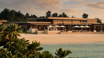 A beachfront hotel with a modern design, featuring a pool area with umbrellas and lounge chairs, and a view of the ocean.