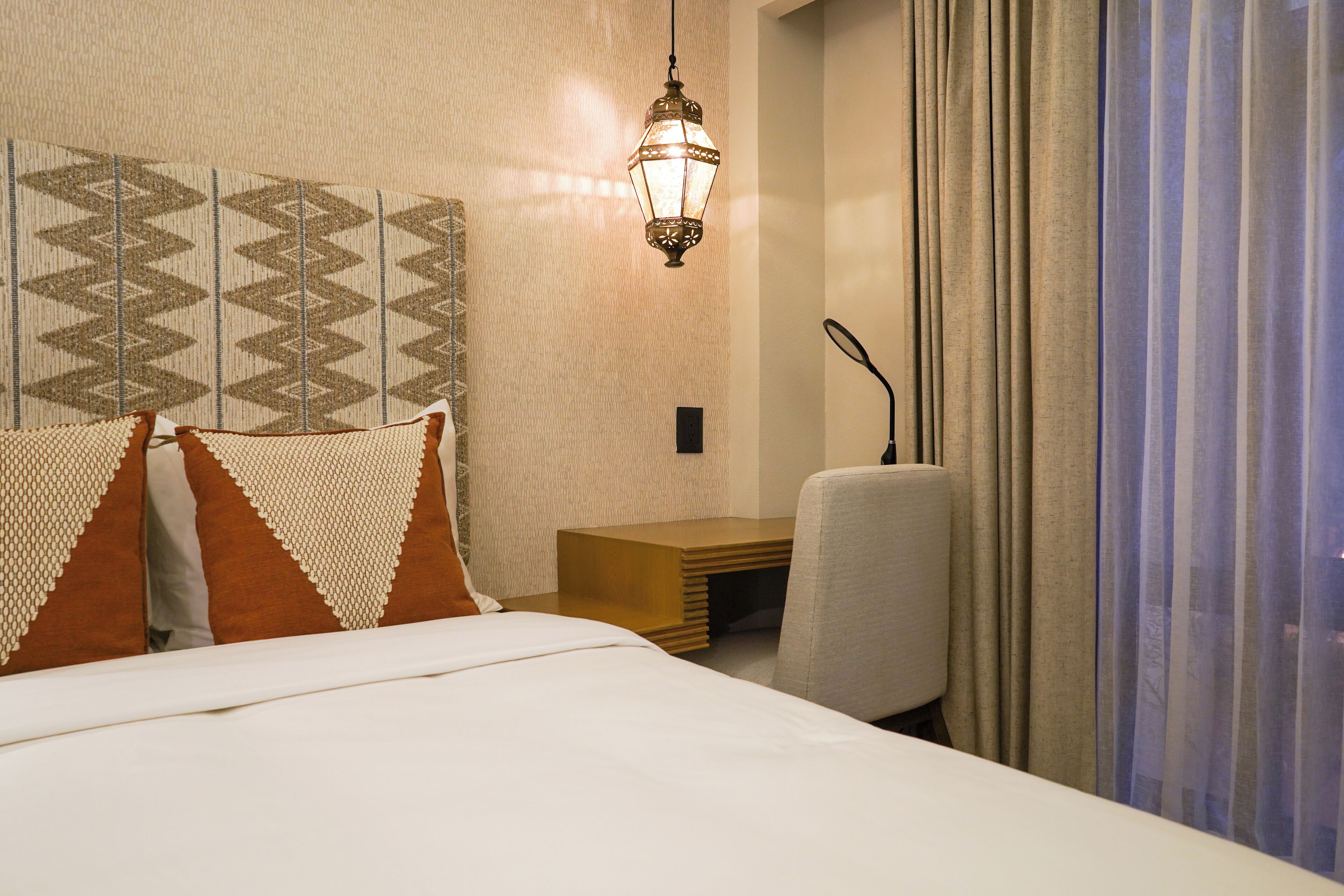 Habitación de hotel con cama, almohadas, escritorio, silla, una lámpara colgante y vistas al exterior.