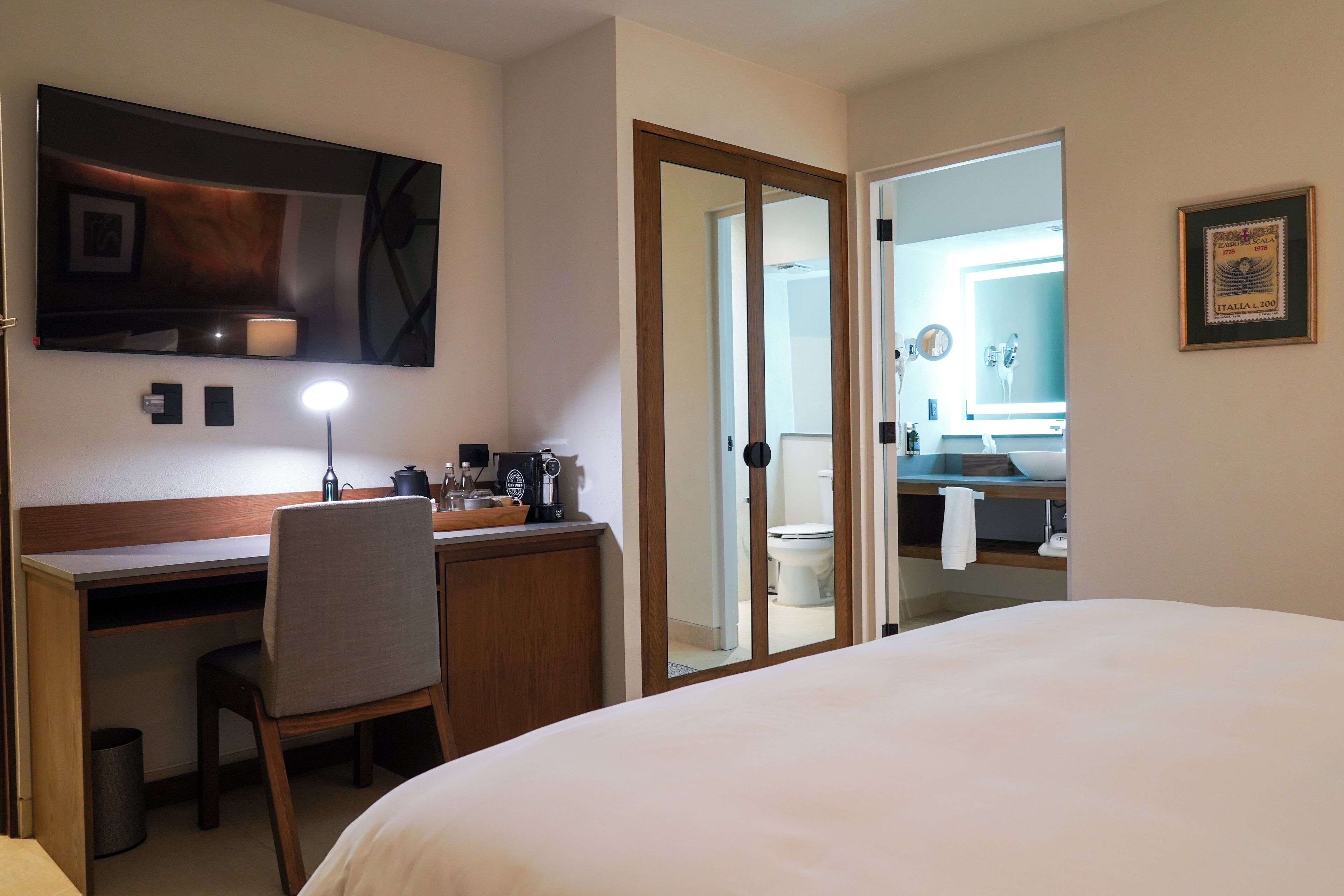 Una habitación de hotel con cama, un escritorio con silla, un espejo, una mesita de noche y un baño visible a través de una puerta abierta.