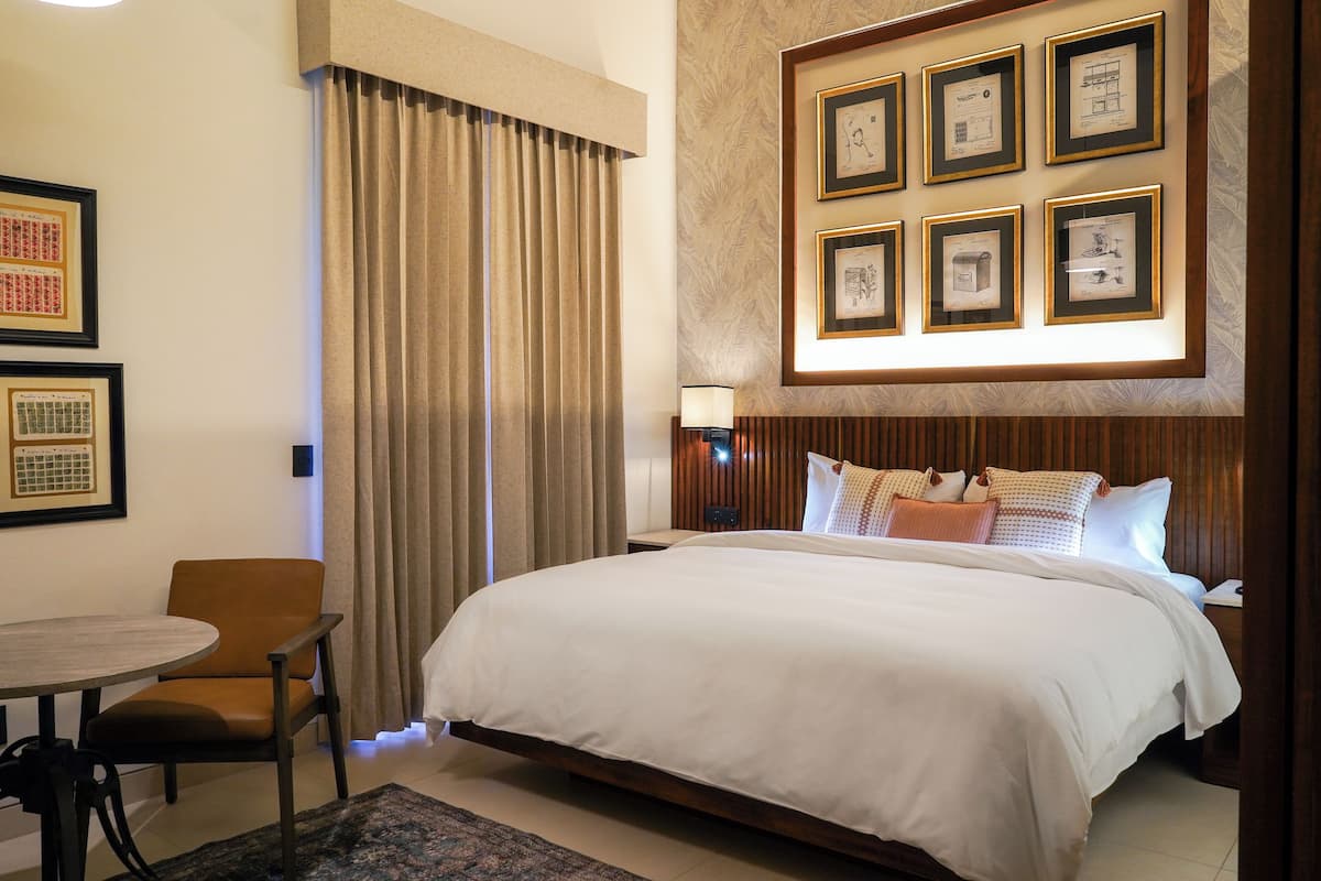 A hotel room with a large bed, a dining table with a chair, and a wall with framed pictures.