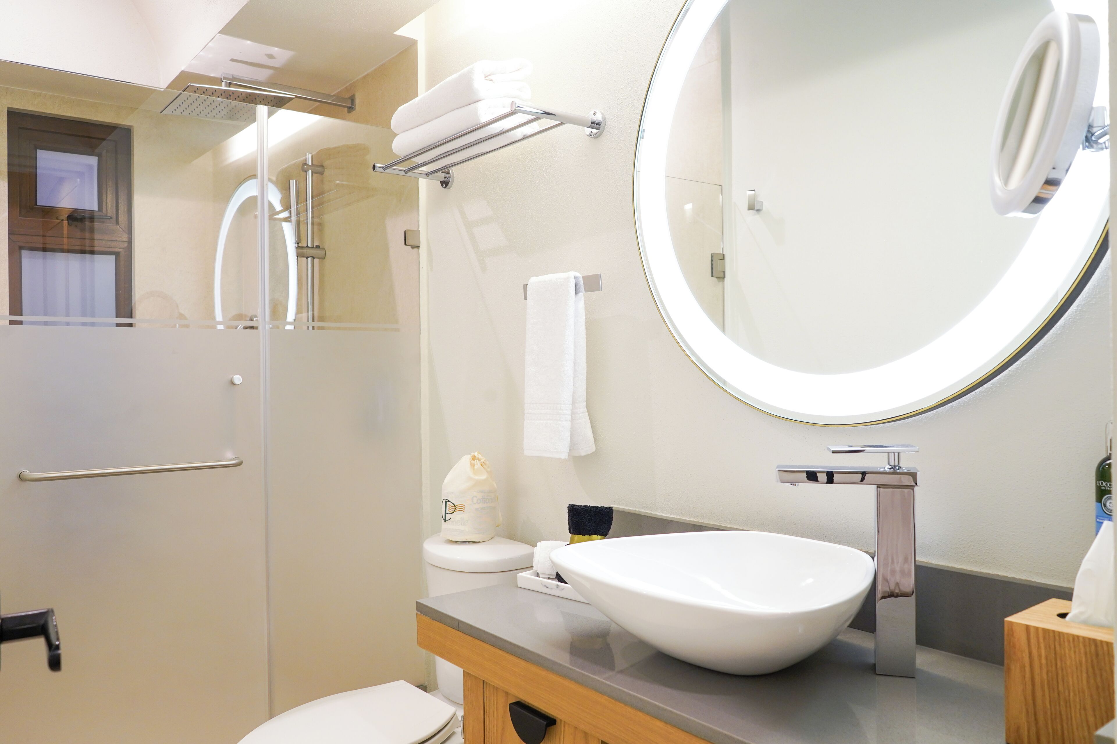 Une salle de bain moderne avec une douche à parois de verre, un lavabo blanc avec un grand miroir et des toilettes surmontées d’un porte-serviettes.