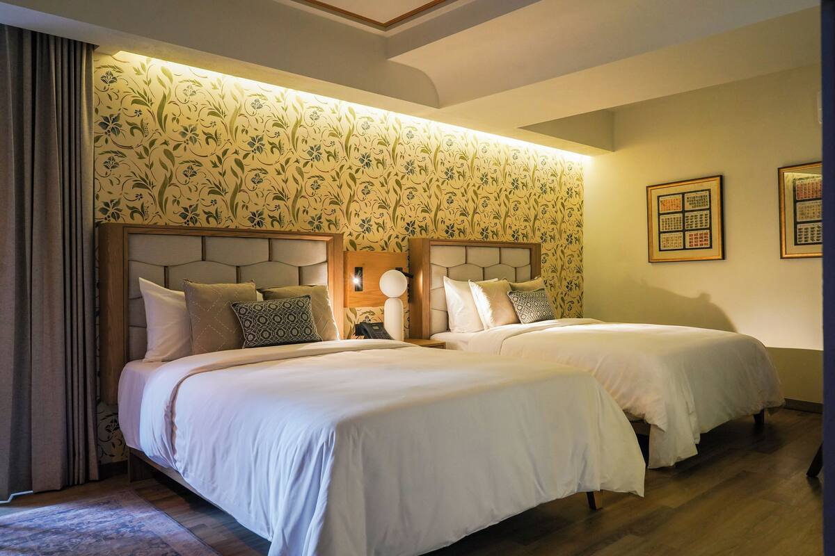 A double room with two beds, a headboard with a light fixture, and a floral wallpaper design.