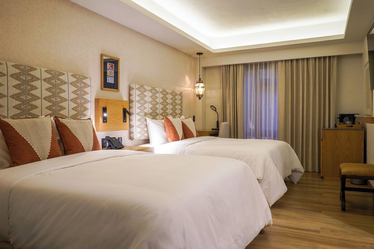 A hotel room with two beds, a desk, a chair, a lamp, and a ceiling light.
