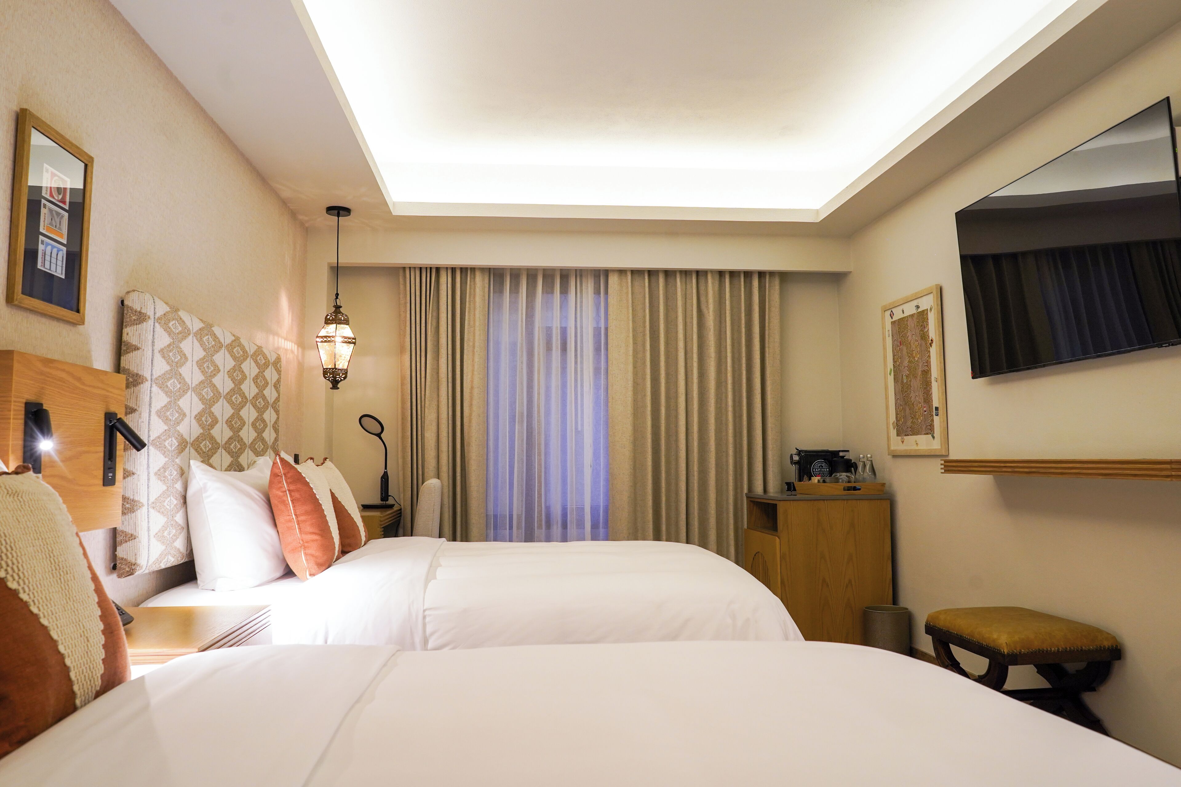 A hotel room with two beds, a desk, a chair, a mirror, a window with curtains, and a television.
