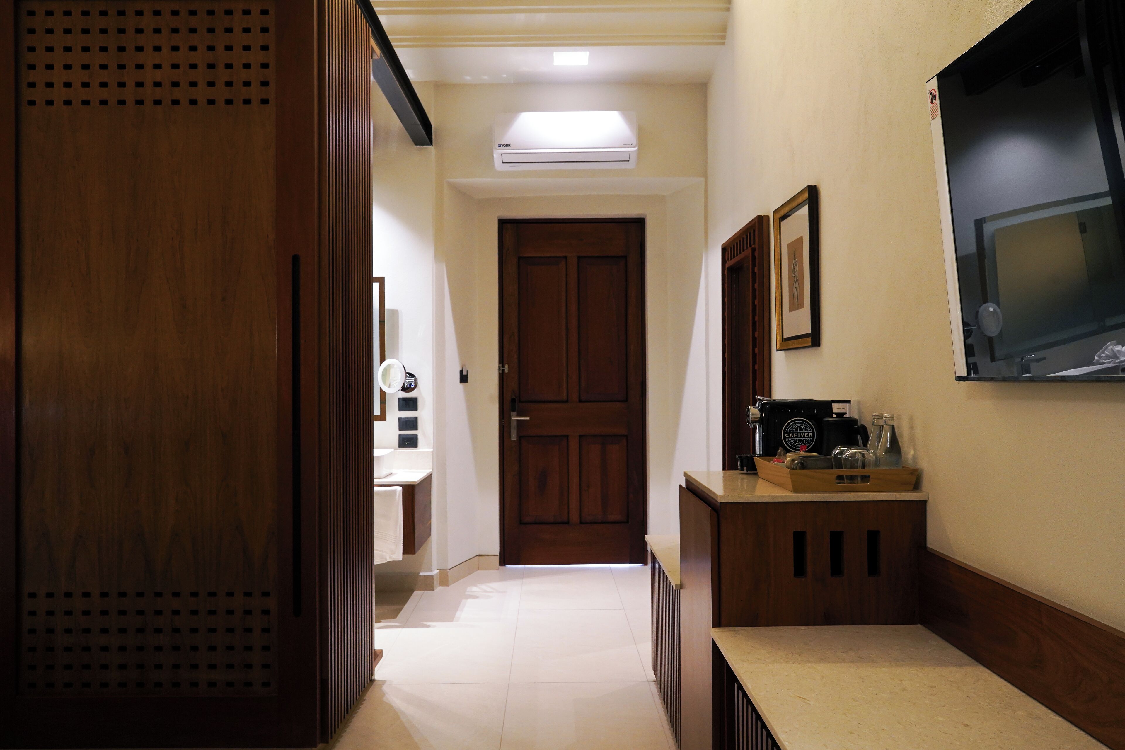 A hallway with a wooden door, a white tiled floor, a small counter with a coffee machine, and a mounted TV.