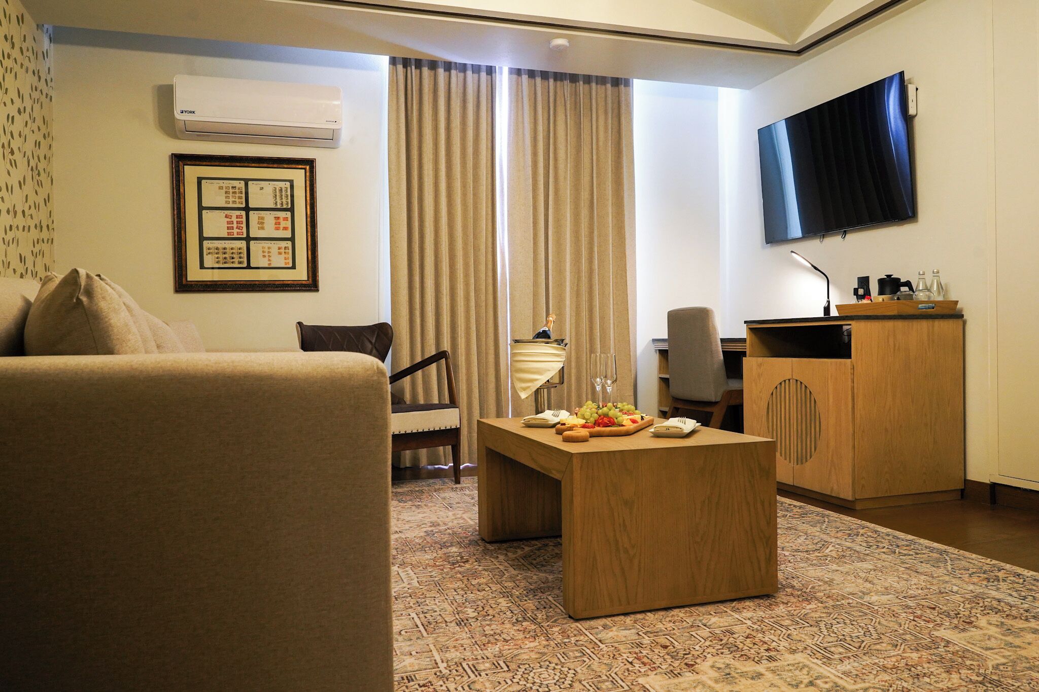 A hotel room with a sofa, a wooden coffee table with fruit, a desk with a computer, a TV, and a patterned rug.