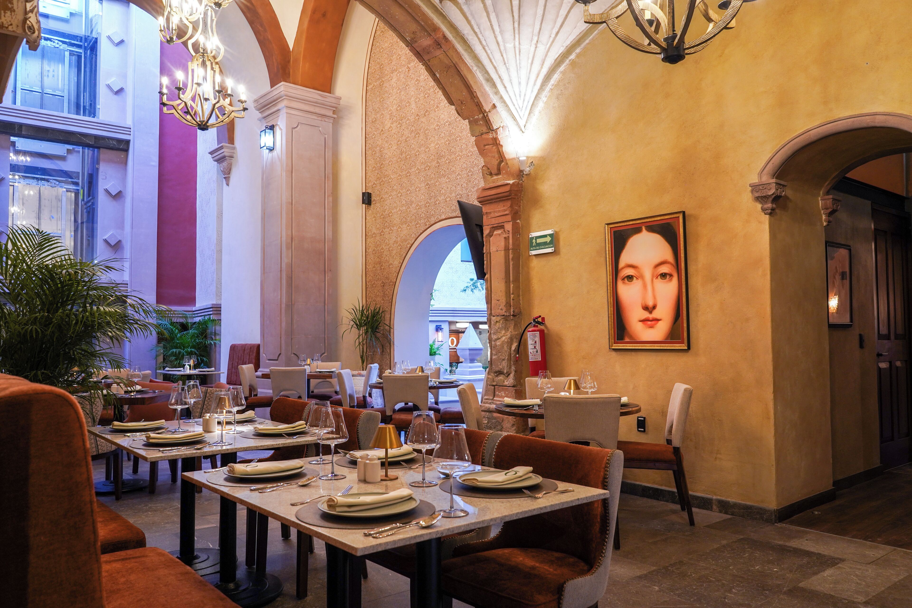 A restaurant with tables set for dining, a chandelier, and a large painting on the wall.
