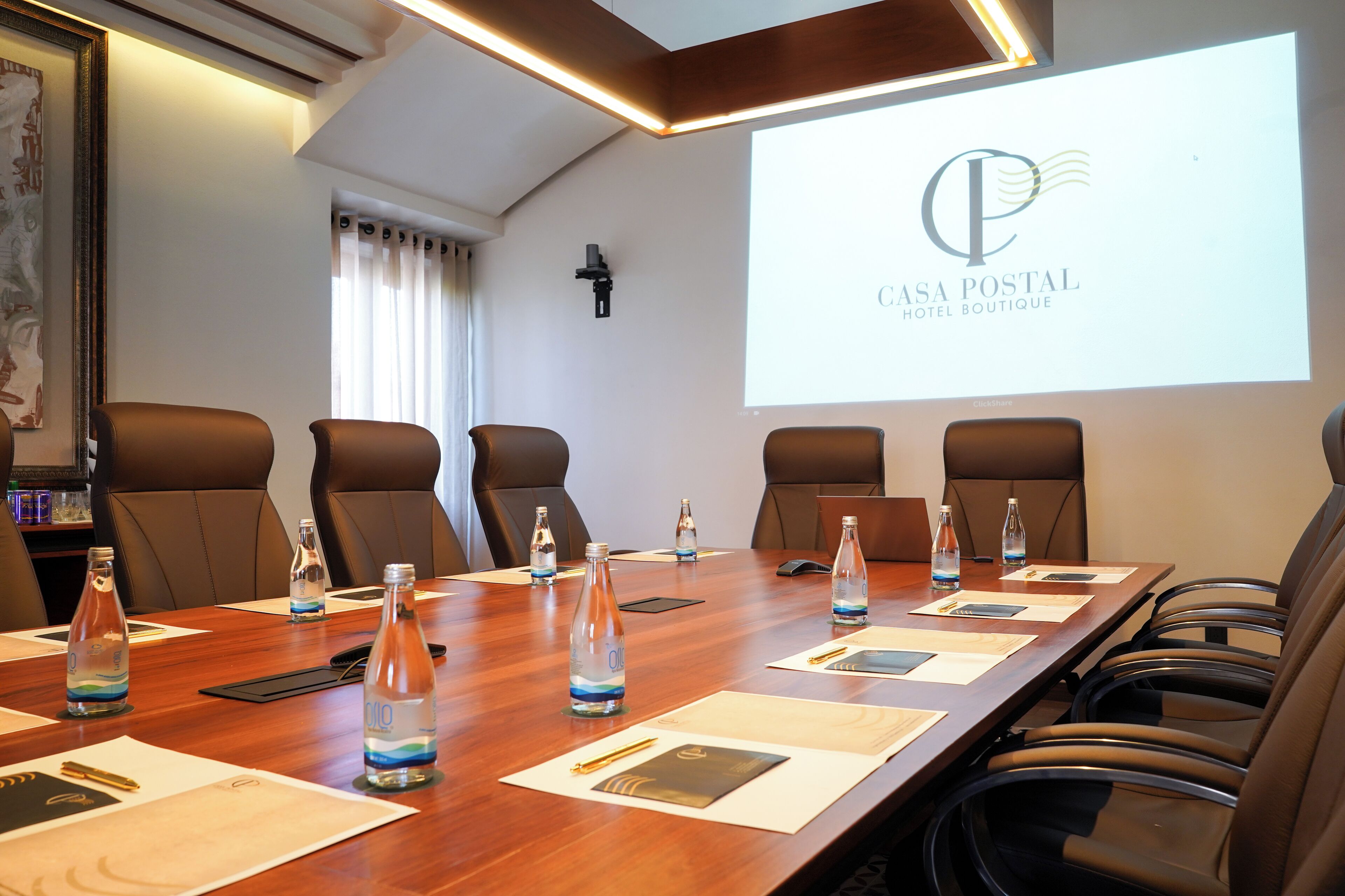 A conference room with a long table, brown leather chairs, water bottles, name cards, and a projector screen displaying "CASA POSTAL HOTEL BOUTIQUE".