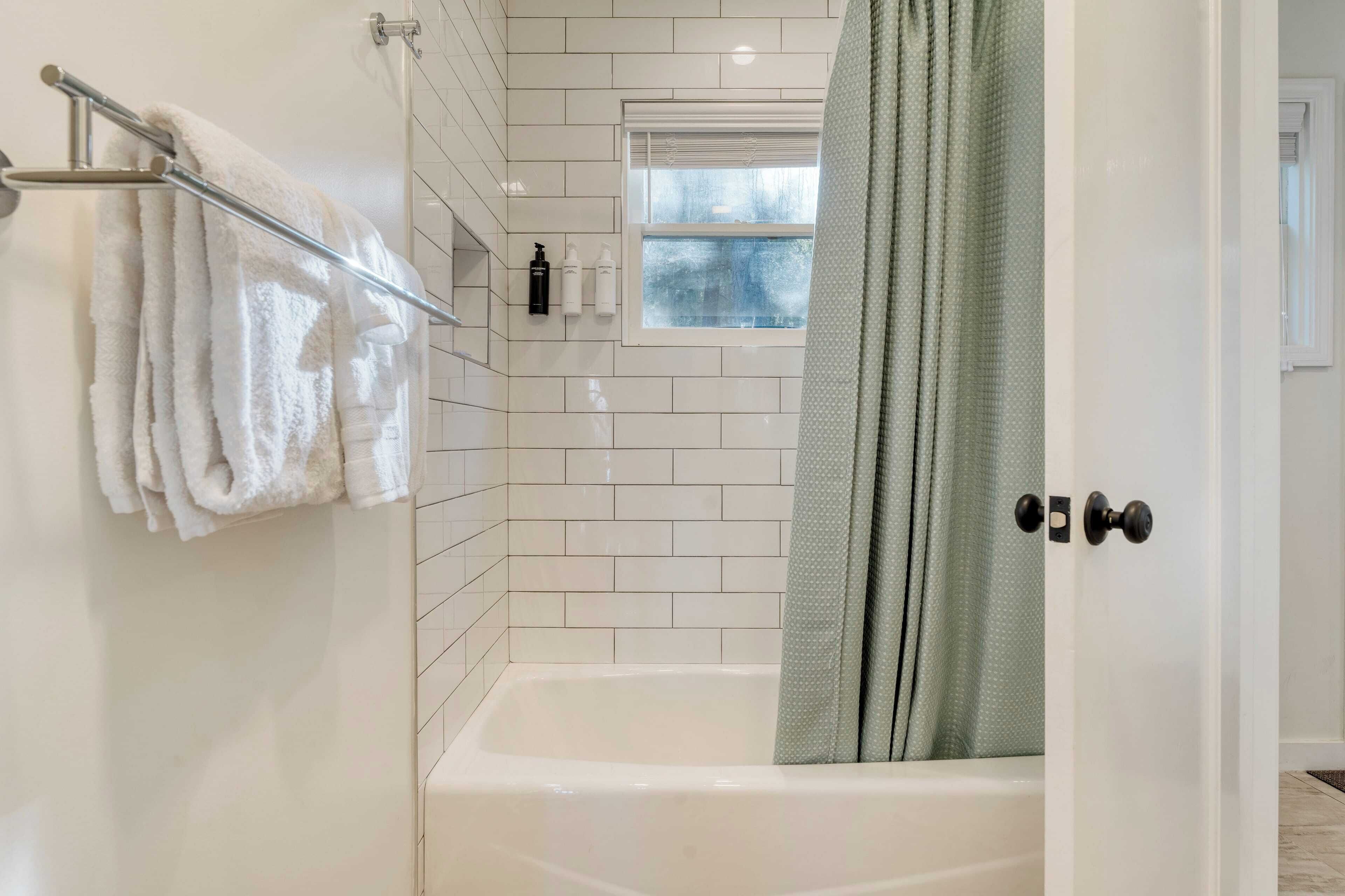 A modern bathroom with white tiles, a bathtub, a towel rack with white towels, and a window with a view.