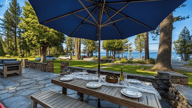 An outdoor dining area with a table set for four, surrounded by stone walls and a large blue umbrella. - West Shore Lake House by Avantstay Ski Cabin w/ Backyard Sledding Hill! (Homewood)
