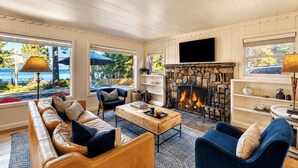 A cozy living room with a stone fireplace, leather sofas, a coffee table, and large windows overlooking a scenic view. - West Shore Lake House by Avantstay Ski Cabin w/ Backyard Sledding Hill! (Homewood)