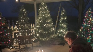 Una persona sentada a una mesa frente a una chimenea decorativa, rodeada de árboles de Navidad con luces.