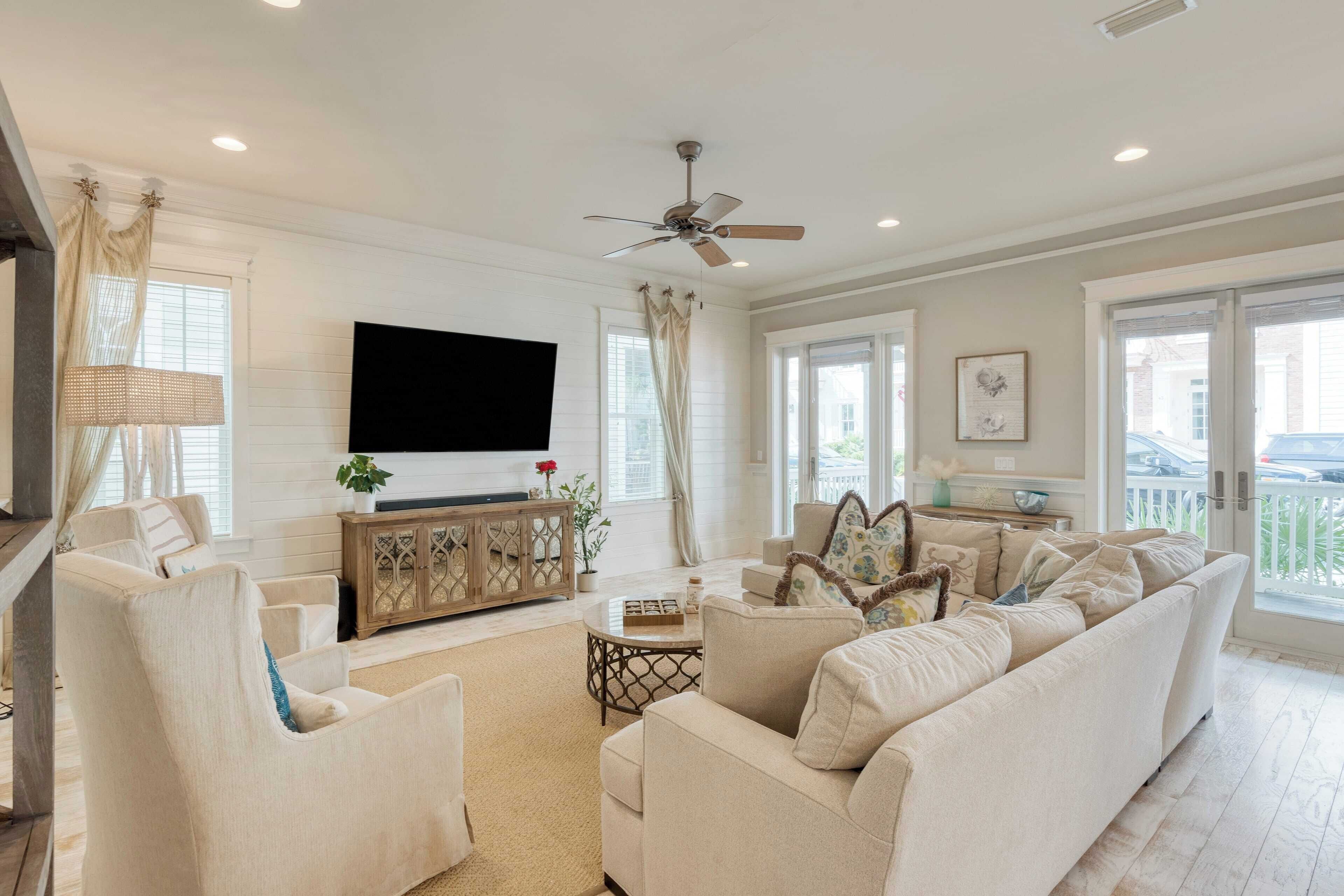 A spacious living room with a large flat-screen TV, comfortable seating, a ceiling fan, and large windows offering a view of the outside.