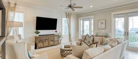 A spacious living room with a large flat-screen TV, comfortable seating, a ceiling fan, and large windows offering a view of the outside.