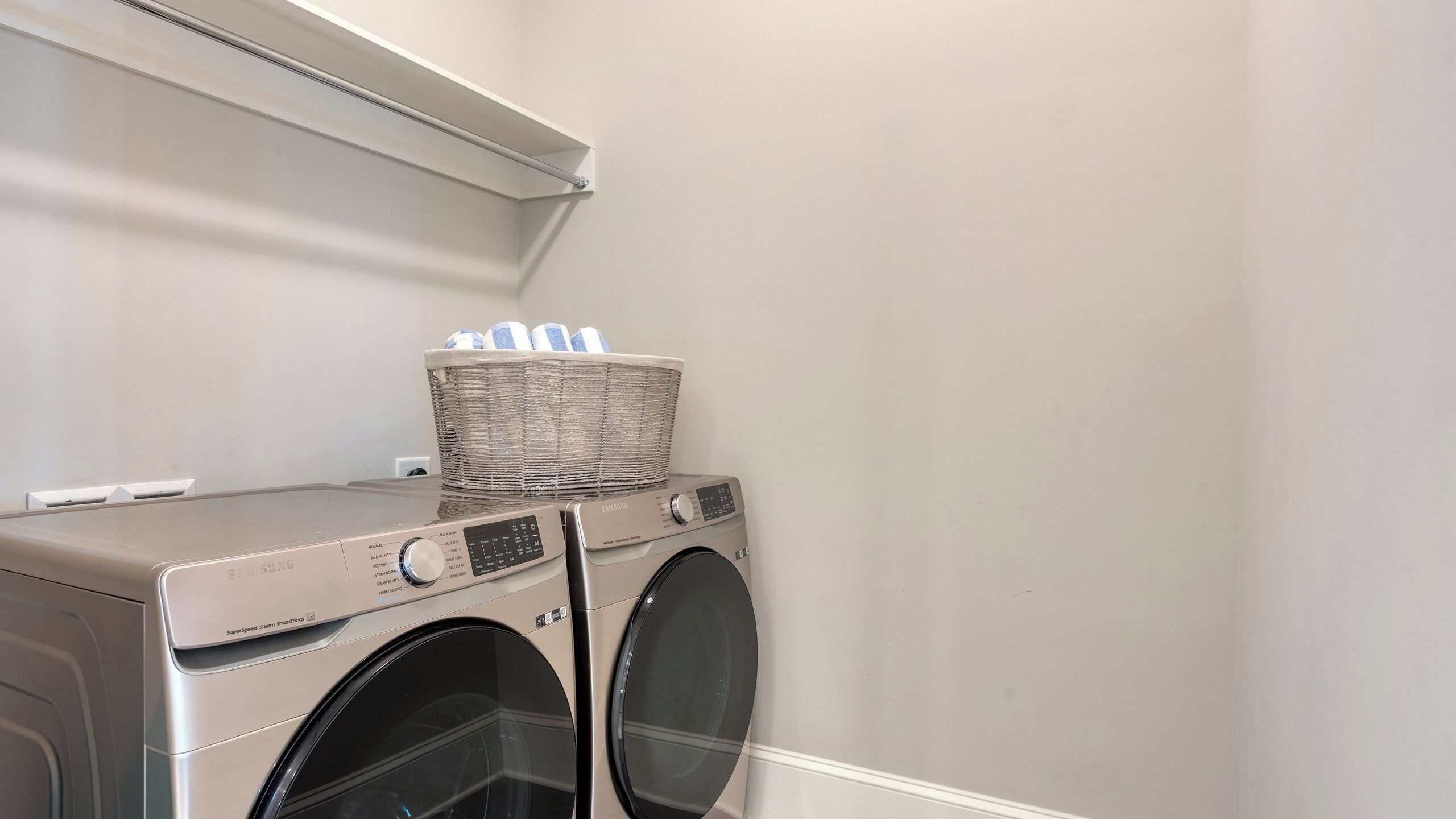 A front-loading washing machine with a basket on top, a wall-mounted shelf above, and a plain wall.
