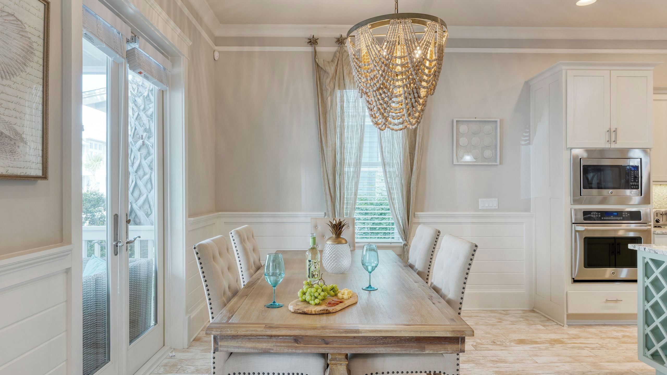 A dining area with a wooden table, cushioned chairs, a chandelier, and a kitchen with stainless steel appliances.