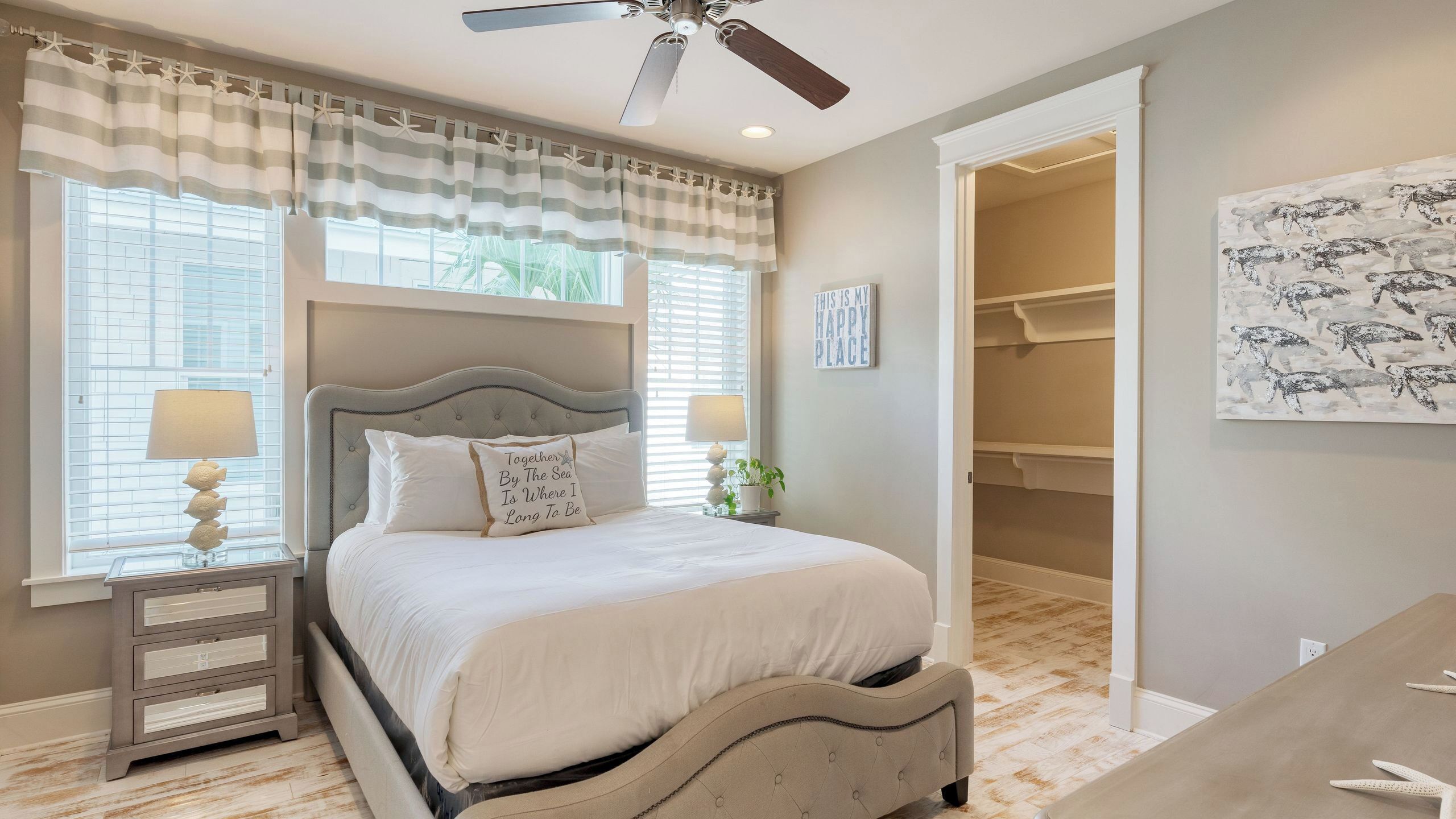 Une chambre à coucher avec un lit, des lampes de chevet, un ventilateur de plafond, une fenêtre avec des rideaux à rayures, une plante sur la table de chevet et un couloir menant à un placard.