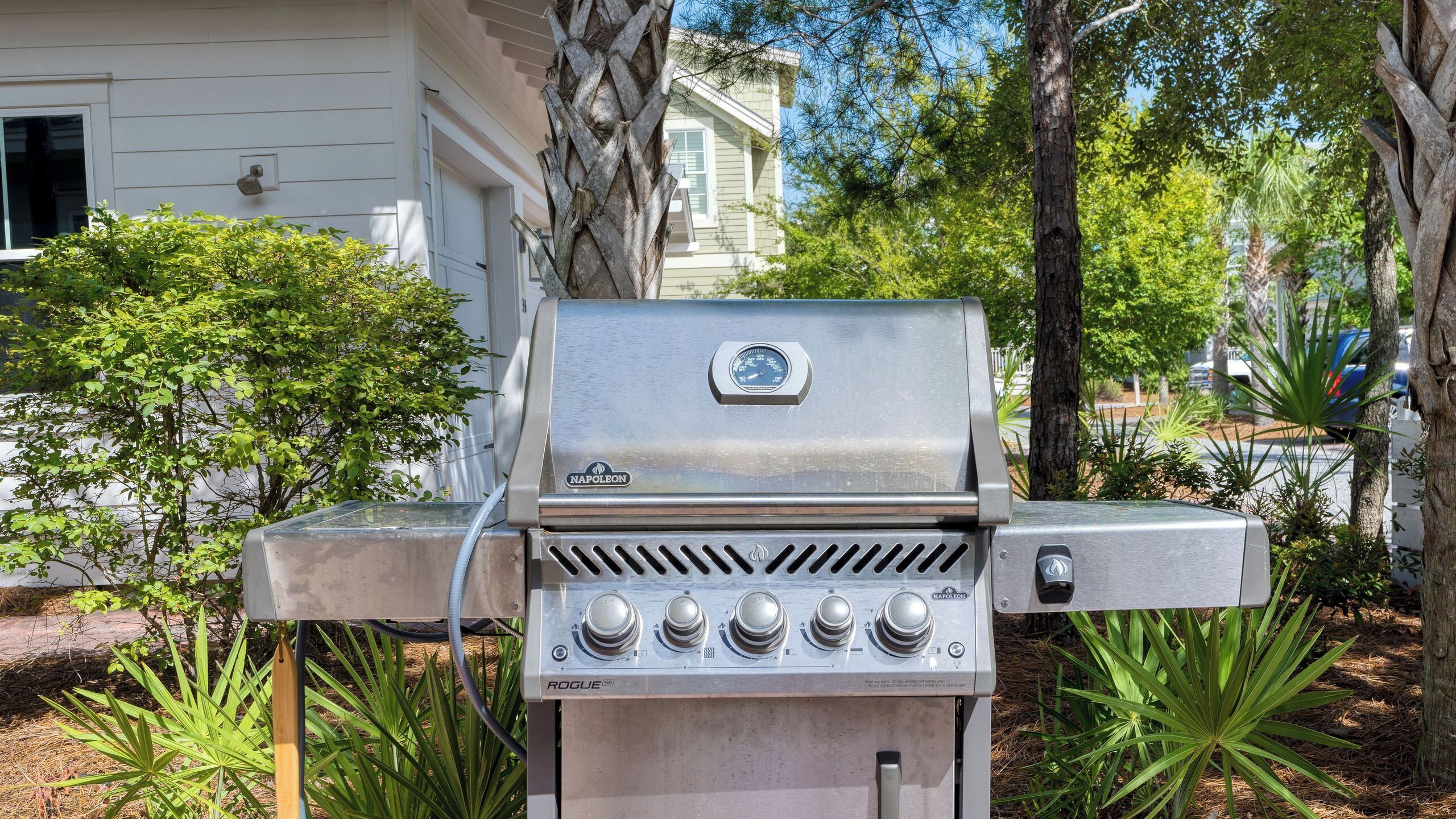 Un barbecue extérieur en acier inoxydable, doté d’un thermomètre numérique, de boutons de contrôle et d’un tuyau d’alimentation en gaz, le tout mis en valeur par un décor d’arbres et d’une maison.