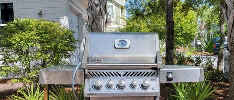 Un barbecue extérieur en acier inoxydable, doté d’un thermomètre numérique, de boutons de contrôle et d’un tuyau d’alimentation en gaz, le tout mis en valeur par un décor d’arbres et d’une maison.