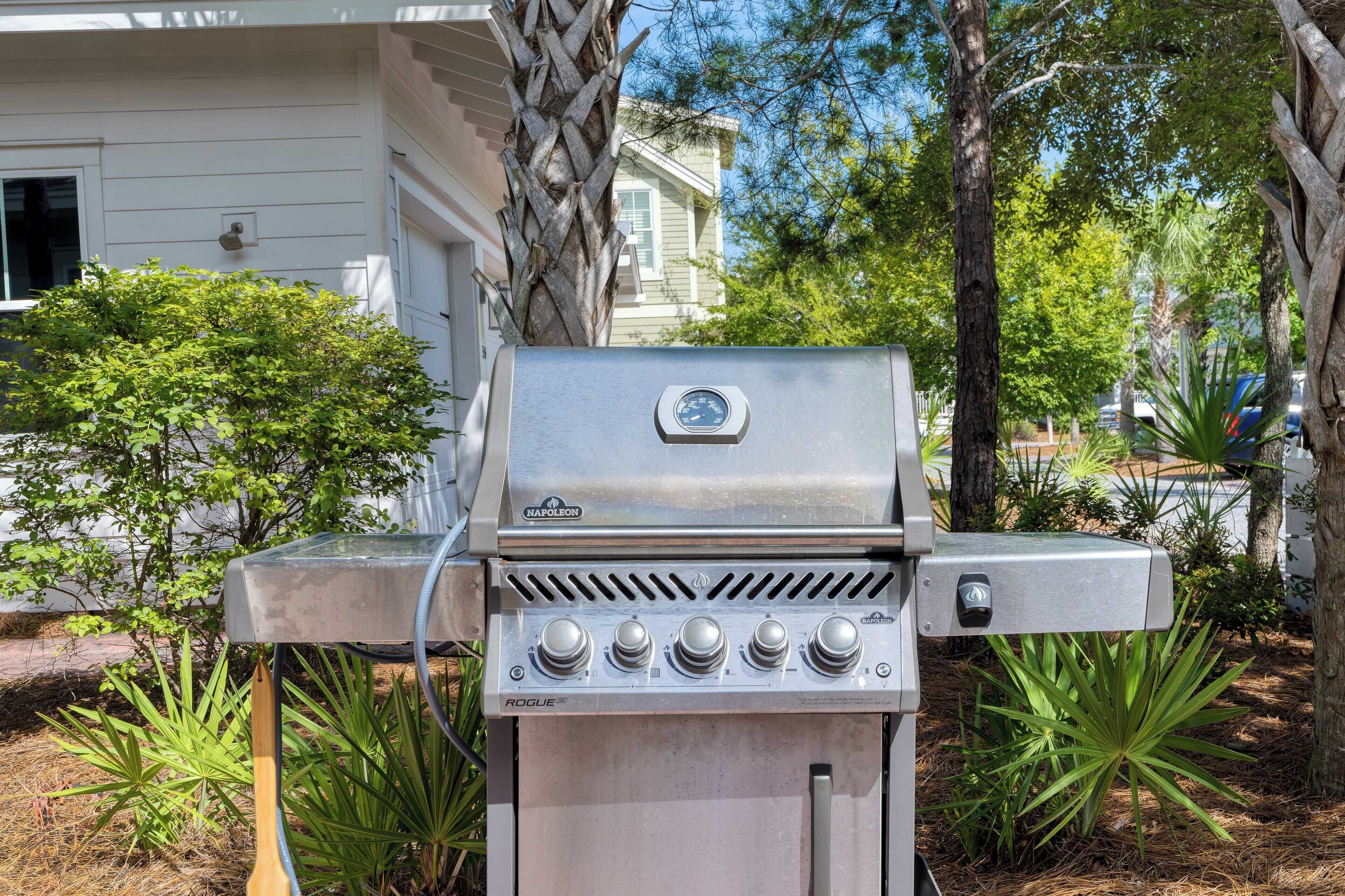 A stainless steel outdoor grill with four control knobs, located in a backyard with trees and a house in the background.