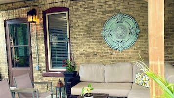 A covered patio area with a brick wall, a cushioned sofa, wicker furniture, and a decorative plate.
