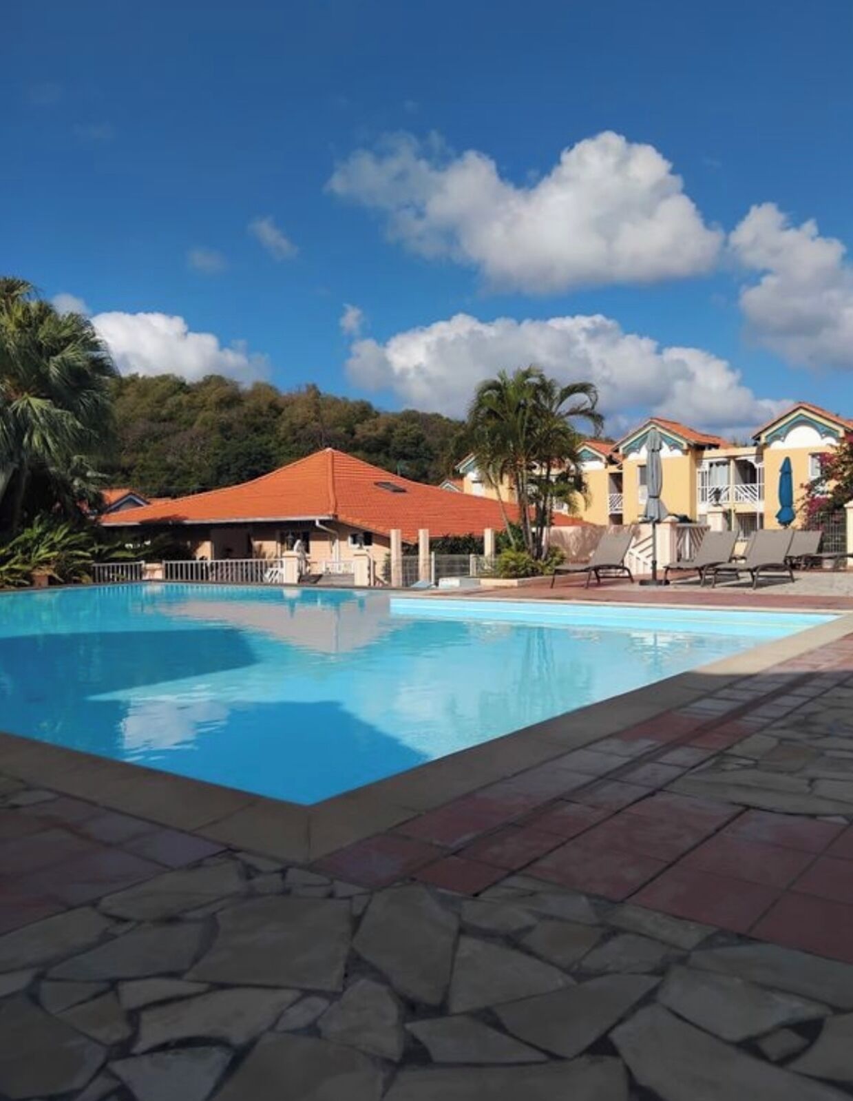 Piscina com espreguiçadeiras e um edifício com telhado vermelho ao fundo.