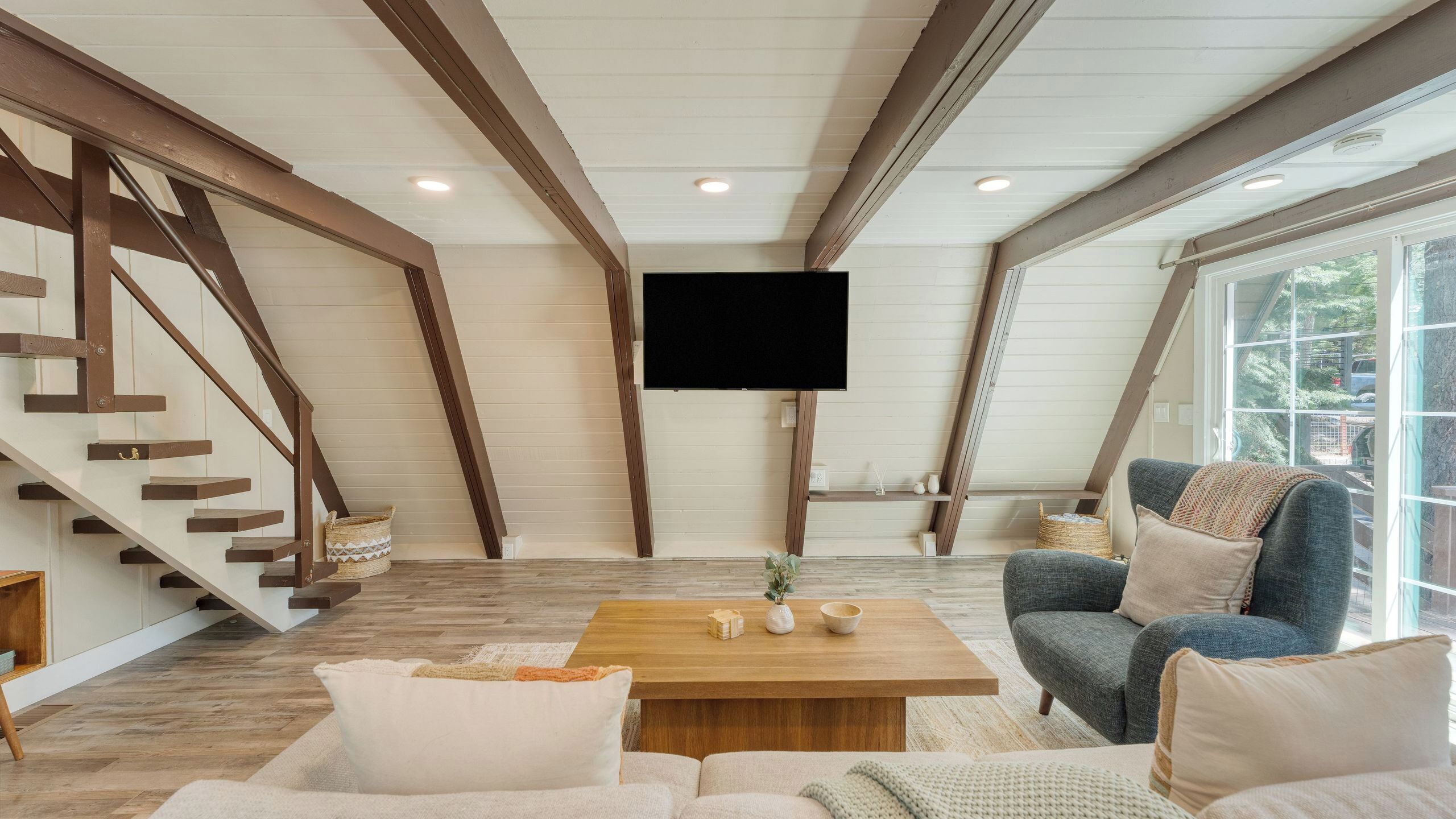 A loft-style living room with exposed wooden beams, a flat-screen TV, a staircase, and large windows.