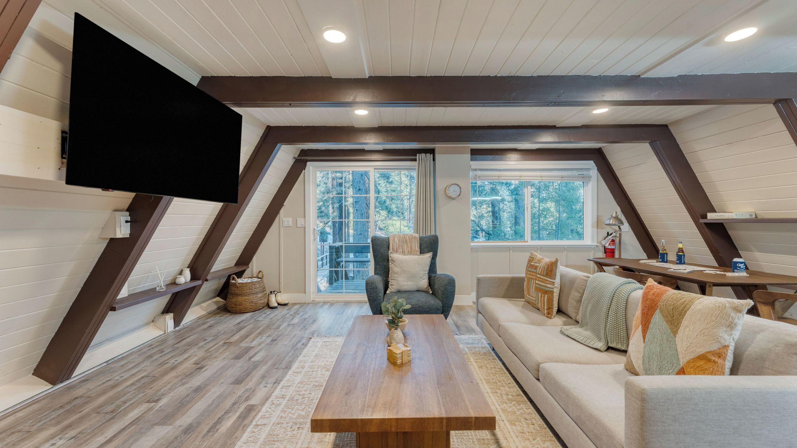 A loft-style room with a wooden floor, a sofa, a coffee table, a TV mounted on the wall, and a window with a view of trees.