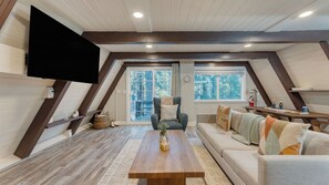 A loft-style room with a wooden floor, a sofa, a coffee table, a TV mounted on the wall, and a window with a view of trees. - Speckled by Avantstay Modern Interior, 0.5mi to Lake! (Kings Beach)
